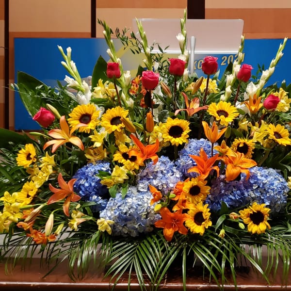 Large stage arrangement with yellow sunflowers, blue hydrangeas, orange lilies, and pink roses