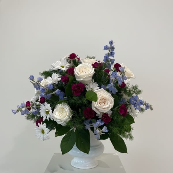 Arrangement of white roses, white daisies, burgundy carnations, and blue delphinium in a white pedestal vase