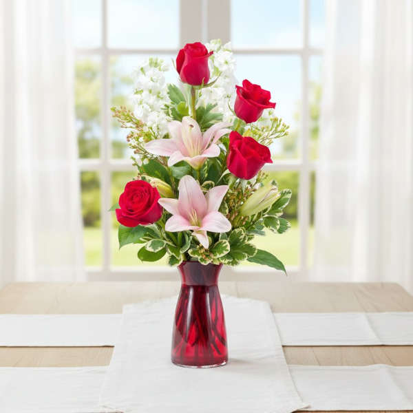Tall arrangement of red roses and pink lilies in a red glass vase on a table