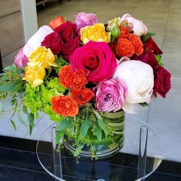 Low glass vase of bright roses, peonies, and spray roses in pink, red, orange, and yellow on a clear stand.