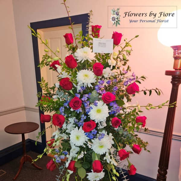 Tall standing spray of red roses, white mums, and blue flowers on a black easel