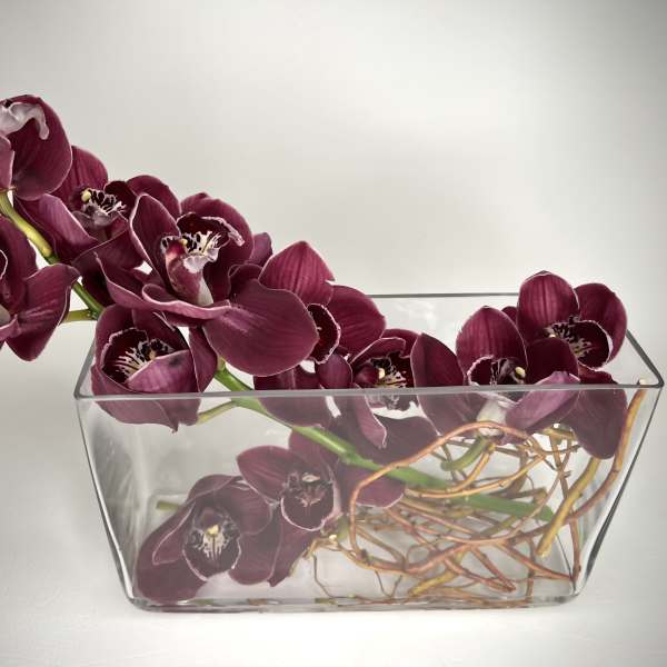 Horizontal arrangement of deep purple cymbidium orchids in a clear rectangular glass vase.