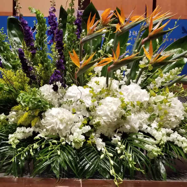 Large stage arrangement with white hydrangeas, orange tropical blooms, and tall green and purple accents