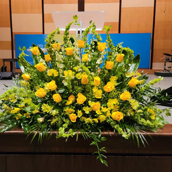 Large fan-shaped arrangement of yellow roses and flowers with lush greenery on a stage front.