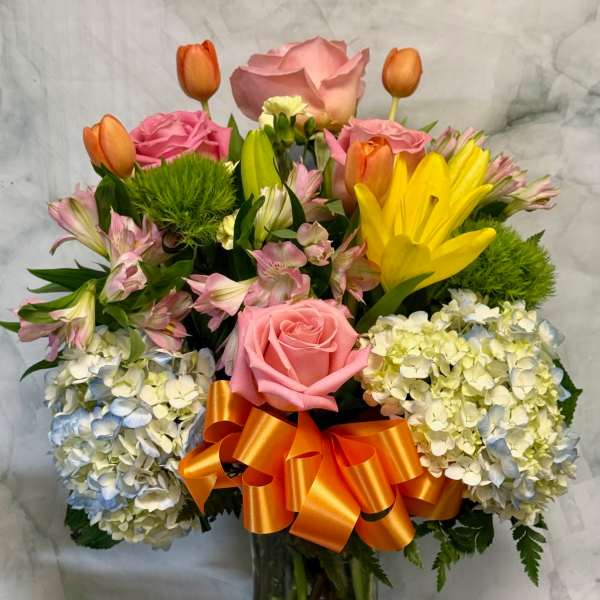 Tall mixed bouquet of pink roses, yellow lilies, tulips and hydrangeas in a clear vase with an orange bow.