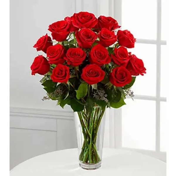 Tall arrangement of long-stemmed red roses in a clear glass vase on a white table.