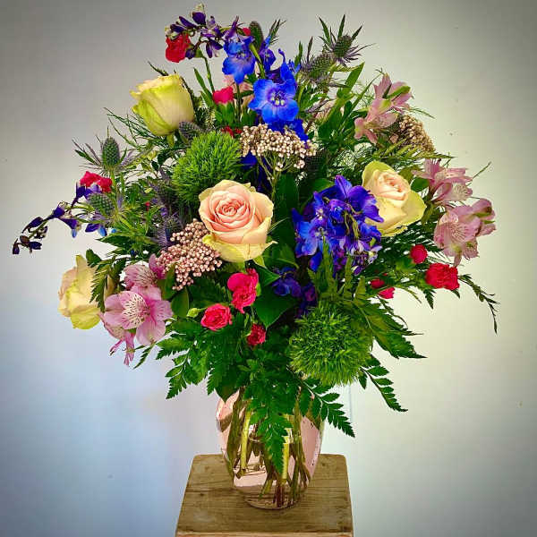 Colorful mixed bouquet with pink roses, blue delphinium, and assorted blooms in a pink glass vase