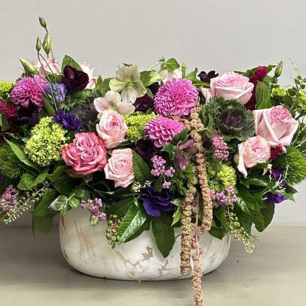 Low mixed arrangement of pink roses, purple blooms, and green accents in a white marble bowl