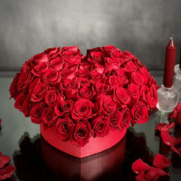 Heart-shaped box of red roses on a reflective surface with scattered petals.