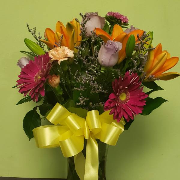 Mixed bouquet of pink gerbera daisies, orange lilies, and roses in a glass vase with a yellow ribbon