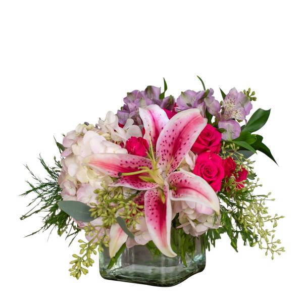 Low arrangement of pink lilies, hot pink roses, hydrangeas, and lavender flowers in a clear glass cube vase