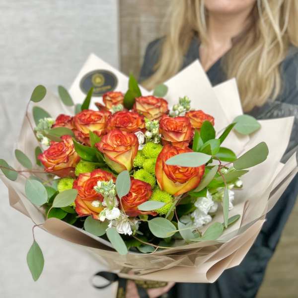 Bouquet of orange and yellow roses with green mums and white blooms wrapped in beige paper