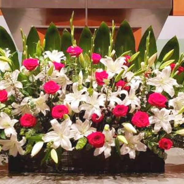 Large low arrangement of red roses and white lilies with bold green leaves on a stage.