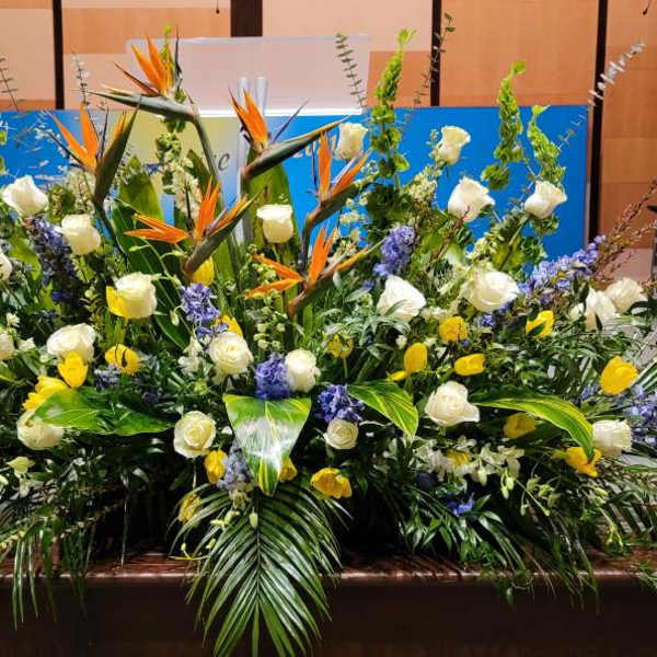 Large stage arrangement of white roses, yellow flowers, blue spikes and orange birds of paradise with lush palm leaves