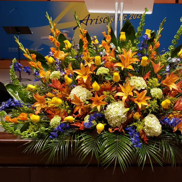 Large altar arrangement with orange lilies, yellow roses, blue delphinium, and white hydrangeas in a fan shape
