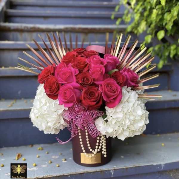 Hatbox arrangement of hot pink and red roses with white hydrangeas, pearls, and metallic fan accents.