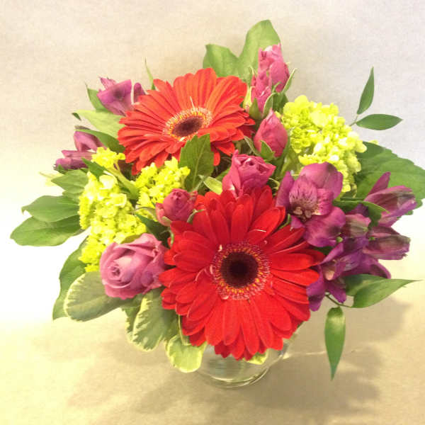 Bouquet of red gerbera daisies, pink roses, and purple flowers in a glass vase