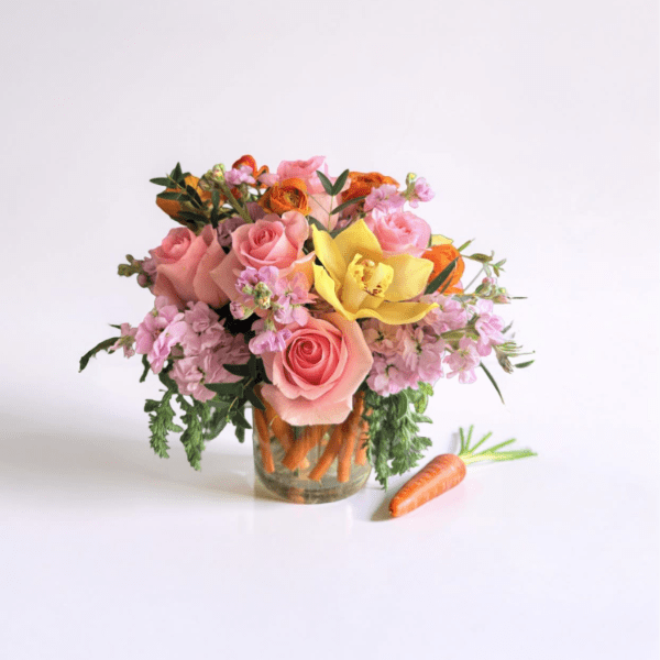 Short arrangement of pink roses, yellow orchid, and orange flowers in a carrot-filled glass vase with a carrot beside it