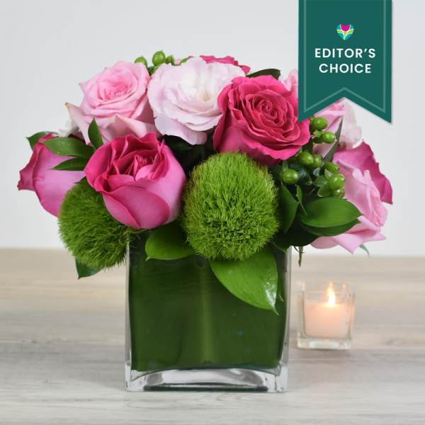 Short arrangement of pink roses and soft pink blooms in a green-lined glass cube vase.