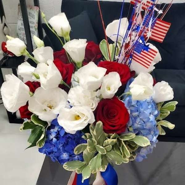 Patriotic red, white, and blue bouquet with roses and hydrangeas in a glass vase with American flags.