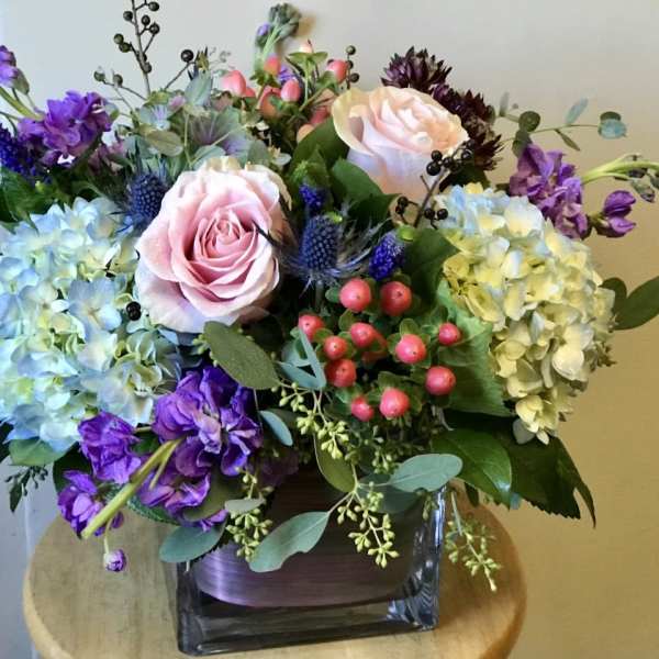 Mixed bouquet with pink roses, blue hydrangeas, and purple flowers in a square vase