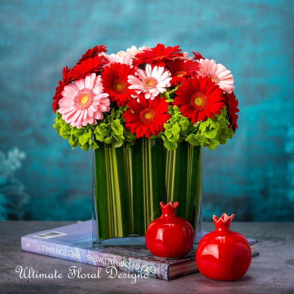 Red and pink gerbera daisies in a clear glass vase with two red pomegranates.