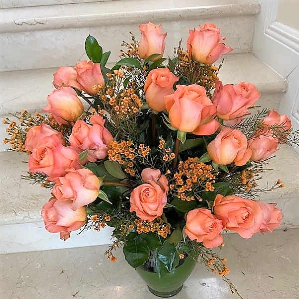 Tall arrangement of peach roses with tiny orange filler blooms in a green glass vase