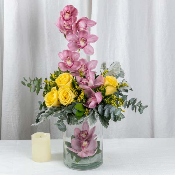 Tall arrangement of pink orchids and yellow roses in a clear glass vase beside a cream pillar candle