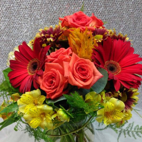 Bouquet of red gerbera daisies, coral roses, and yellow alstroemeria in a glass vase
