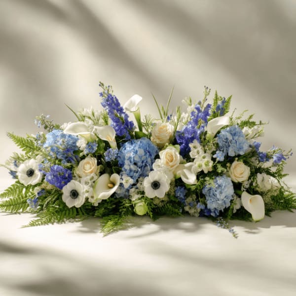 Low arrangement of blue hydrangeas, white calla lilies, roses, and anemones in a horizontal spray