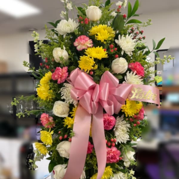 Standing spray of pink, yellow, and white flowers with a large pink bow on an easel.