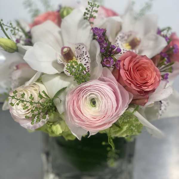 Short arrangement of pink ranunculus, roses, white orchids, and purple blooms in a clear glass cube vase