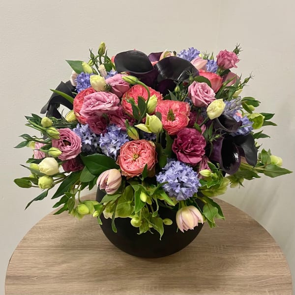 Low black bowl arrangement of pink roses, purple calla lilies, lisianthus, and lilac hyacinths