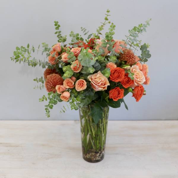Tall glass vase arrangement of orange and peach roses with dahlias and eucalyptus.