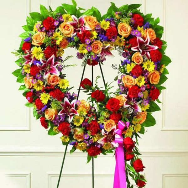 Heart-shaped standing wreath of red, yellow, orange, and purple flowers with a pink ribbon.