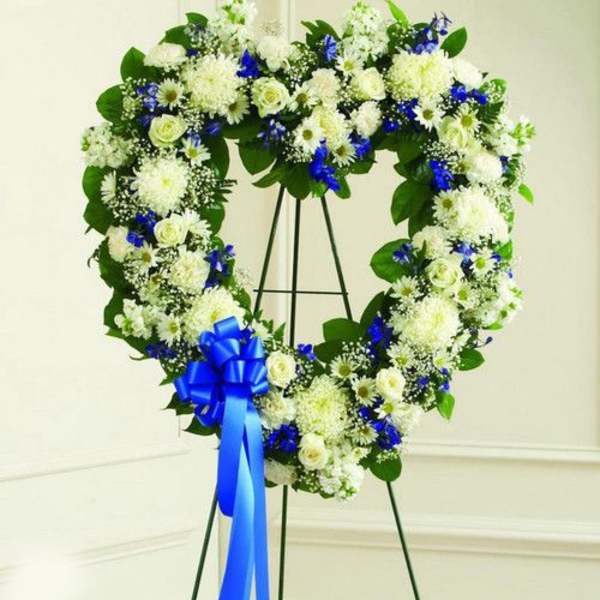 Heart-shaped white and blue funeral wreath on a stand with a blue bow.