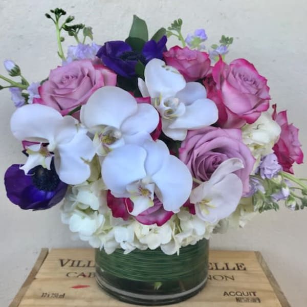 Round arrangement of white orchids, hydrangeas, and pink and purple roses in a glass vase.