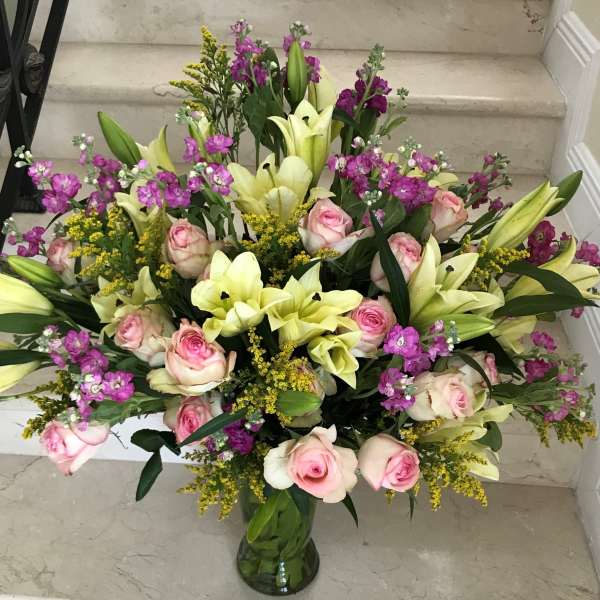 Large mixed bouquet of pale yellow lilies, pink roses, and purple flowers in a clear glass vase