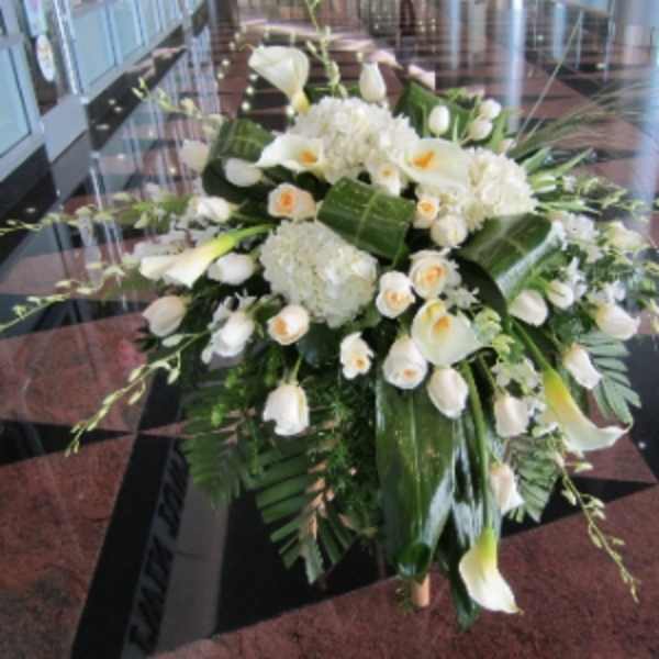 Large white arrangement of roses, calla lilies, and hydrangeas with bold green leaves on a stand.