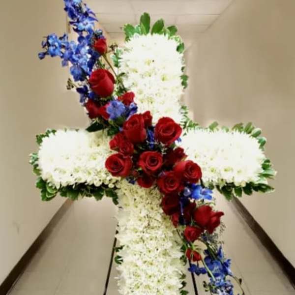 Tall white floral cross with red roses and blue flowers on a metal stand