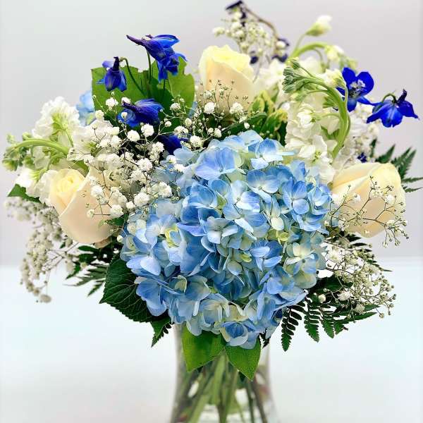 Vase arrangement with blue hydrangeas, cream roses, white blooms, and bright blue flowers