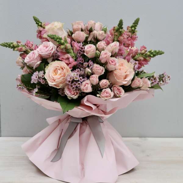 Hand-tied bouquet of light pink roses and snapdragons wrapped in pink paper with gray ribbon