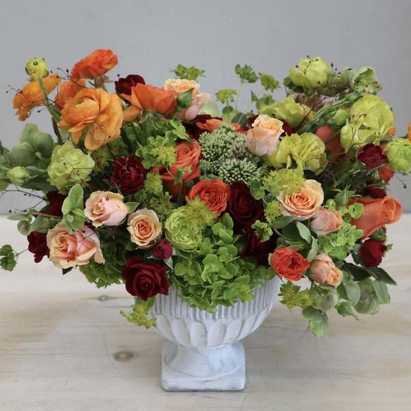 Lush arrangement of orange, peach, red, and green flowers in a white pedestal vase