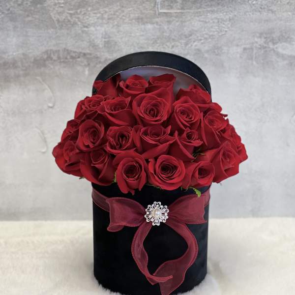 Round black hatbox filled with red roses, tied with a red bow and jeweled brooch.
