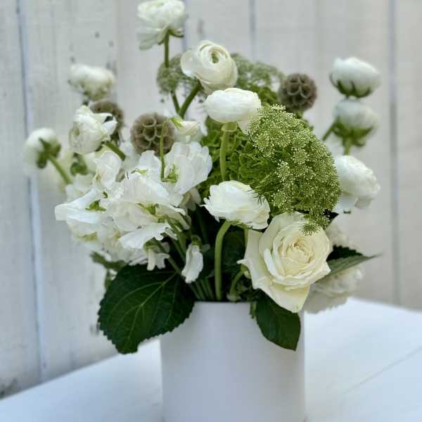 White roses and ranunculus with green accent blooms arranged in a white cylinder vase.