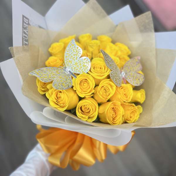 Handheld bouquet of bright yellow roses with silver butterfly accents wrapped in white and kraft paper.