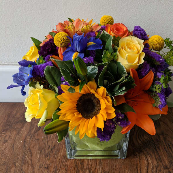 Colorful bouquet with sunflowers, roses, lilies, and purple filler in a square glass vase