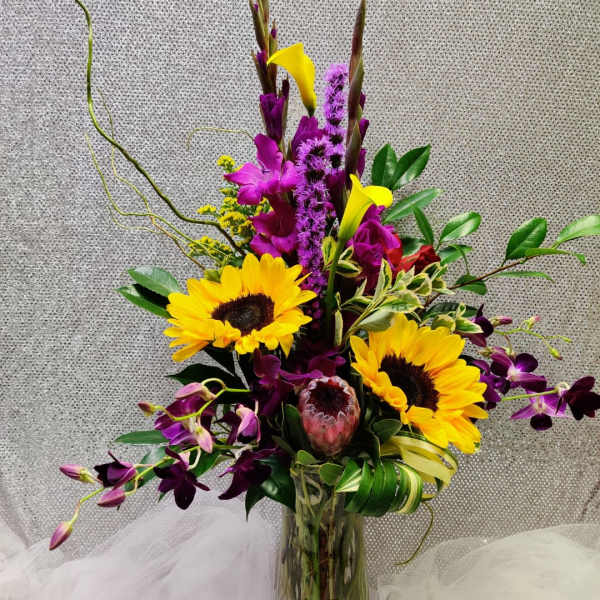 Tall mixed arrangement with sunflowers, purple orchids, yellow calla lilies, and protea in a clear glass vase.