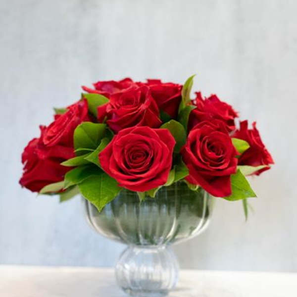 Low arrangement of red roses in a clear pedestal glass vase on a light surface