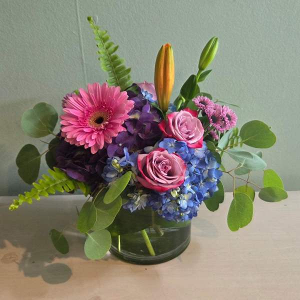 Pink gerbera, roses, blue hydrangea, and lilies in a glass vase